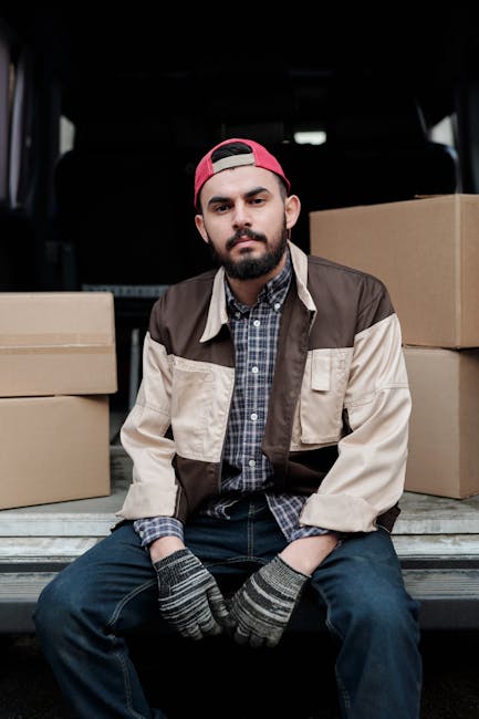 A young man with a beard, wearing a black cap and a white polo shirt, stands inside the rear of a moving van with its doors open. Behind him, several cardboard boxes of various sizes are stacked neatl