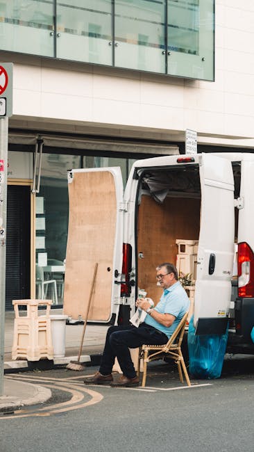 A man wearing glasses and a light blue shirt is seated on a wooden chair next to an open white moving van parked on a city street in front of a modern building with large glass windows. Inside the van, visible wooden furniture pieces and cardboard boxes are prepared for home relocation or furniture transport. The man is holding a document or map, possibly reviewing moving details. Nearby, a wooden board and a small plastic crate are positioned on the pavement. The scene captures the loading process where boxes, packaging materials, and furniture are being organized for transportation, indicative of packing and moving activities associated with professional removals services. The environment suggests a busy urban setting, with natural lighting illuminating the scene, supporting the context of house removals and logistics handled by companies like Man with Van Twickenham.
