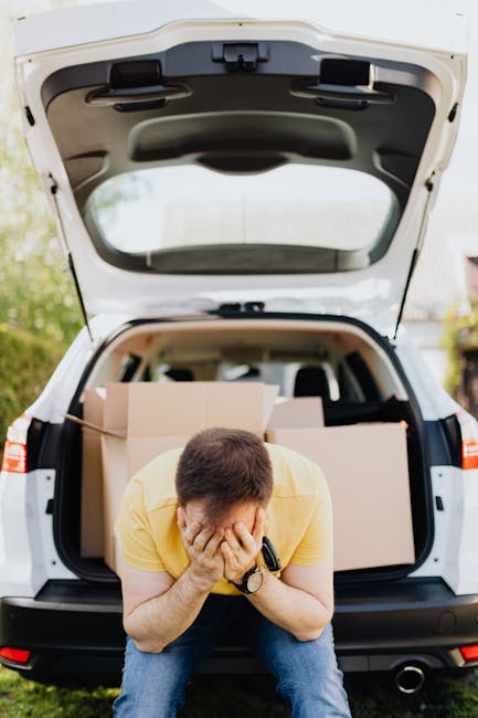A man wearing a yellow shirt and blue jeans is sitting on the pavement in front of an open car trunk, covering his face with his hands. Inside the trunk, several large cardboard boxes and packing materials are visible, indicating a home relocation or moving process. The vehicle is parked outdoors in a residential area with greenery and houses in the background. The scene captures a moment during furniture transport or packing and moving activities, which are part of house removals services. The image reflects the logistical aspects of a house move, as handled by companies such as Man with Van Twickenham, including loading and transportation of household items.